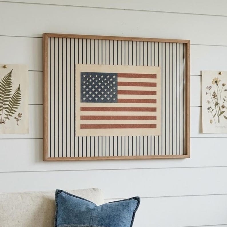 Puede incluir: Una pieza de arte mural rectangular enmarcada que presenta una impresi&oacute;n centrada de la bandera de los Estados Unidos con una paleta de colores rojo, blanco y azul apagados. La bandera se sit&uacute;a sobre un fondo de rayas verticales negras en papel crema.