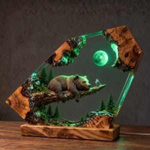 May include: A decorative lamp featuring a brown bear resting on a tree branch within a clear resin and natural wood frame. The scene includes miniature pine trees, a small bird, and a glowing green moon, all set upon a rectangular wooden base.