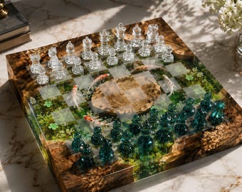 Op de afbeelding: Een vierkant schaakbord van hout en hars met een vijverontwerp met ingebedde koikarpers, groen gebladerte en een centraal steenachtig element. Het bord bevat een set doorzichtige glazen schaakstukken en een set donkergroenblauwe glazen schaakstukken.