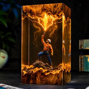 May include: A rectangular resin lamp featuring a miniature figure of a worker in an orange high-visibility shirt and blue trousers standing on a rocky base. The figure holds a glowing, lightning-like electrical discharge reaching toward the top.