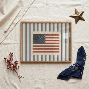 Puede incluir: Un marco de madera rectangular que contiene una impresi&oacute;n de la bandera estadounidense centrada sobre un fondo de finas rayas verticales azules. La bandera presenta un cant&oacute;n azul con estrellas blancas y franjas horizontales rojas y blancas.