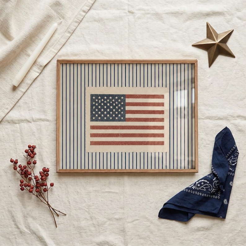 Puede incluir: Un marco de madera rectangular que contiene una impresi&oacute;n de la bandera estadounidense centrada sobre un fondo de finas rayas verticales azules. La bandera presenta un cant&oacute;n azul con estrellas blancas y franjas horizontales rojas y blancas.