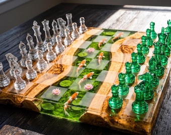 Op de afbeelding: Een schaakspel met een bord van natuurlijk hout en groene hars met ingebedde koi-vissen en lotusbloemmotieven. De set bevat heldere glazen schaakstukken aan de ene kant en doorschijnende groene glazen schaakstukken aan de andere kant.
