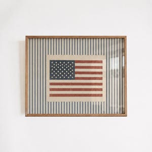 Puede incluir: Una impresi&oacute;n rectangular enmarcada que presenta una ilustraci&oacute;n centrada de la bandera de los Estados Unidos con una textura desgastada. La bandera se sit&uacute;a sobre un fondo de rayas verticales azul marino sobre un campo de color crema.