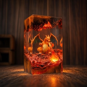May include: A rectangular resin lamp featuring an orange dragon-like creature with wings, positioned inside a clear block. The base and top contain textured, dark red and orange material resembling lava, with a glowing light effect.