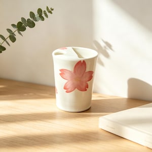 Könnte beinhalten: Ein weißer Keramikbecher mit einem Design aus rosa Kirschblüten. Der Becher hat eine einzigartige Form mit einer geschwungenen Oberseite und einer kleinen Öffnung. Das florale Design wiederholt sich um den Becher. Er steht auf einer Holzoberfläche.