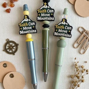 May include: Three pens with the text "Faith Can Move Mountains" on the top. The pens are in shades of blue, green, and olive. The image also includes small brown stones, paper tags, and a compass.