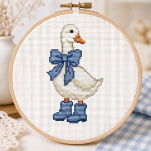 May include: Cross-stitch embroidery of a white goose wearing a blue bow and blue boots, set in a wooden embroidery hoop. The goose has an orange beak and black eyes. The background is white fabric.