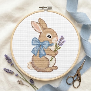 May include: Cross-stitch embroidery of a brown bunny with a blue bow, holding lavender sprigs. The bunny is in a wooden embroidery hoop. Light blue ribbon and small scissors are also in the image.