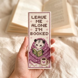 Pode incluir: Um marcador de livros bordado com a frase "LEAVE ME ALONE I'M BOOKED". O marcador apresenta um simpático gambá lendo um livro sob um cobertor roxo, com uma caneca de café. Um livro aberto está no fundo.