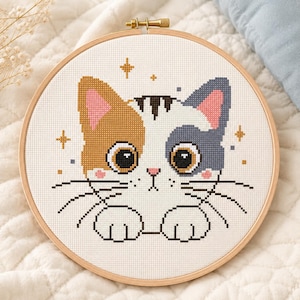 Op de afbeelding: Een kruissteek borduurproject in een ronde houten borduurring met een lapjeskat met grote bruine ogen, roze wangen en een klein roze neusje. De kat heeft één oranje oor en één grijs oor, met witte vacht en zwarte snorharen, omringd door gouden sterren.