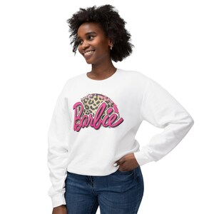 May include: White sweatshirt featuring the word "Barbie" in pink script with a leopard print design. The sweatshirt has long sleeves and a crew neck. The design is centered on the chest.