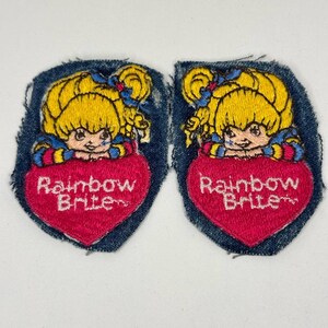 May include: Two embroidered fabric patches featuring the character Rainbow Brite with yellow hair and blue bows, resting her chin on a large pink heart. The text Rainbow Brite is stitched in white across the centre of each heart on a denim background.