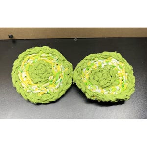 May include: Two circular coasters crafted from braided fabric strips in shades of lime green, yellow, and white. The coasters feature a coiled, textured design with a dense, woven construction that creates a layered, multi-coloured appearance.