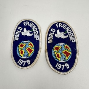 May include: Two oval-shaped embroidered patches with a dark blue background. Each patch features a white dove holding a green olive branch above a globe with yellow and red continents. The text reads World Friendship and 1979 in white thread.