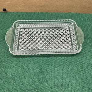 May include: A clear glass rectangular serving tray featuring a pressed diamond pattern across the centre and a decorative scalloped edge. The tray includes two integrated handles on the short sides, providing a functional design for serving.
