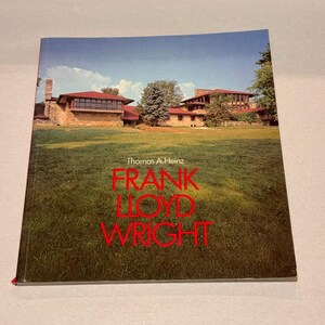 May include: A hardcover book titled Frank Lloyd Wright by Thomas A. Heinz. The cover features a photograph of a stone house with a low-pitched roof set against a green lawn and blue sky. The title is printed in large, bold red capital letters.