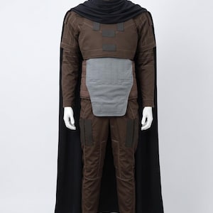 May include: A brown long-sleeved shirt and matching trousers set featuring a grey abdominal plate, dark brown rectangular shoulder and leg patches, and a black cape draped over the shoulders with a high collar, displayed on a white mannequin.