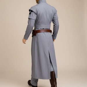 May include: A light gray costume with a high collar, long sleeves, and a long coat. The costume includes a brown belt and boots. The mannequin has a gold head. The costume is a sci-fi or fantasy style.
