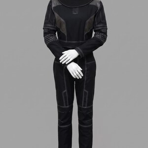 K&ouml;nnte beinhalten: Ein schwarzer Langarm-Jumpsuit mit hohem Stehkragen, strukturierten Schulterpartien und wei&szlig;en Kontrastn&auml;hten. Das Kleidungsst&uuml;ck verf&uuml;gt &uuml;ber texturierte geometrische Muster an den Unterarmen und ein k&ouml;rperbetontes Design.