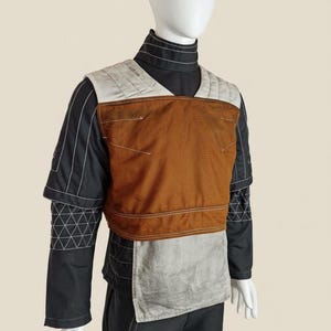 May include: A costume featuring a black long-sleeved shirt with white stitching and a brown and grey vest. The vest has a quilted design on the shoulders and a rectangular panel in the centre. The costume is displayed on a mannequin.