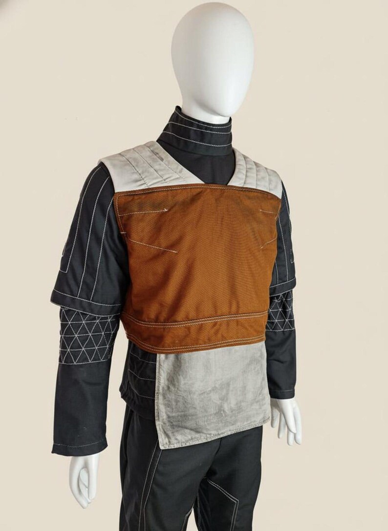 May include: A costume featuring a black long-sleeved shirt with white stitching and a brown and grey vest. The vest has a quilted design on the shoulders and a rectangular panel in the centre. The costume is displayed on a mannequin.