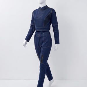 Op de afbeelding: Een donkerblauwe tweedelige outfit bestaande uit een cropped top met lange mouwen en een hoge kraag en een bijpassende broek. De set is voorzien van opvallende witte stiksels, gestructureerde panelen en donkerblauwe accenten.