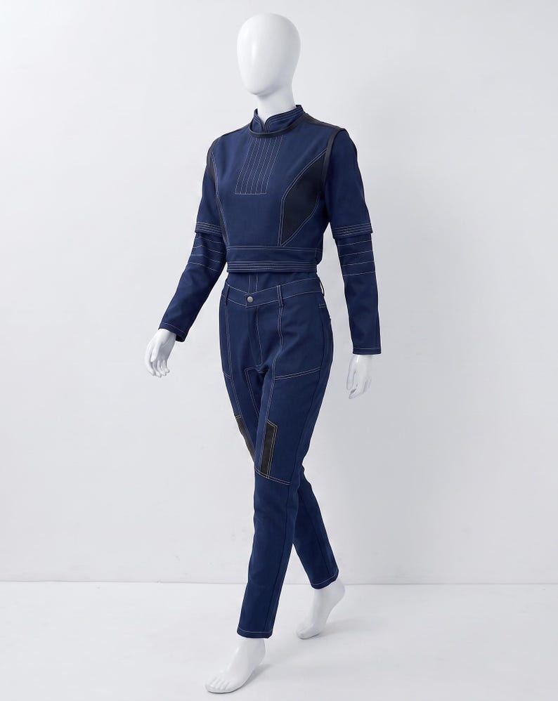 Op de afbeelding: Een donkerblauwe tweedelige outfit bestaande uit een cropped top met lange mouwen en een hoge kraag en een bijpassende broek. De set is voorzien van opvallende witte stiksels, gestructureerde panelen en donkerblauwe accenten.