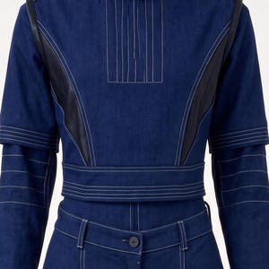Op de afbeelding: Een donkerblauwe spijkerstof top en bijpassende broek met een hoge kraag met zwarte bies, witte contraststiksels en zwarte zijpanelen in lederlook. De top heeft korte mouwen over lange mouwen en de broek heeft een knoopsluiting.
