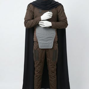 May include: A brown long-sleeved shirt and matching trousers set featuring a black cape, a high black collar, and a grey chest plate. The outfit includes dark brown rectangular patches on the shoulders and thighs, with visible stitching details.
