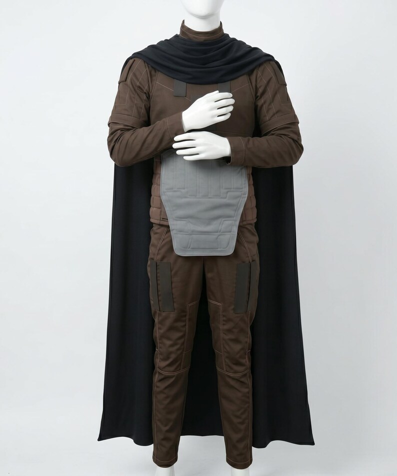 May include: A brown long-sleeved shirt and matching trousers set featuring a black cape, a high black collar, and a grey chest plate. The outfit includes dark brown rectangular patches on the shoulders and thighs, with visible stitching details.