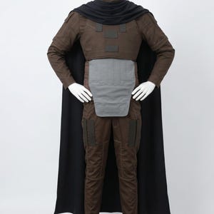 May include: A brown long-sleeved tactical costume featuring a black cape and cowl, a grey abdominal plate, and dark brown rectangular accents on the chest and thighs. The outfit includes fitted trousers and a structured top with a high neck collar.