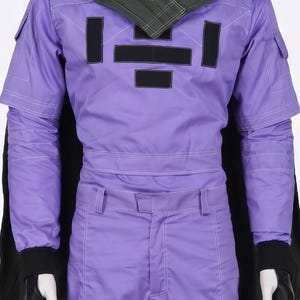 May include: A purple long-sleeved jumpsuit featuring a dark green cape, black rectangular patches on the chest and legs, and black fingerless gloves. The garment includes white topstitching details, a high collar, and utility-style pockets.
