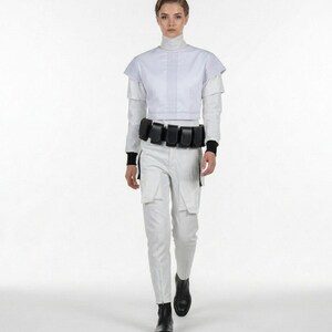 Pu&ograve; includere: Un outfit di ispirazione futuristica con un top corto bianco con cuciture verticali, maniche a strati e pantaloni bianchi abbinati. Una cintura nera con pi&ugrave; tasche e stivali neri completano il look. La modella sta camminando.