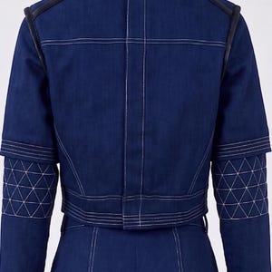 Op de afbeelding: Een donkerblauw spijkerjack met een opstaande kraag, zwarte lederachtige biezen op de schouders en witte contraststiksels. De mouwen hebben een gewatteerd driehoekspatroon en het jack is gecombineerd met een bijpassende donkerblauwe spijkerbroek.