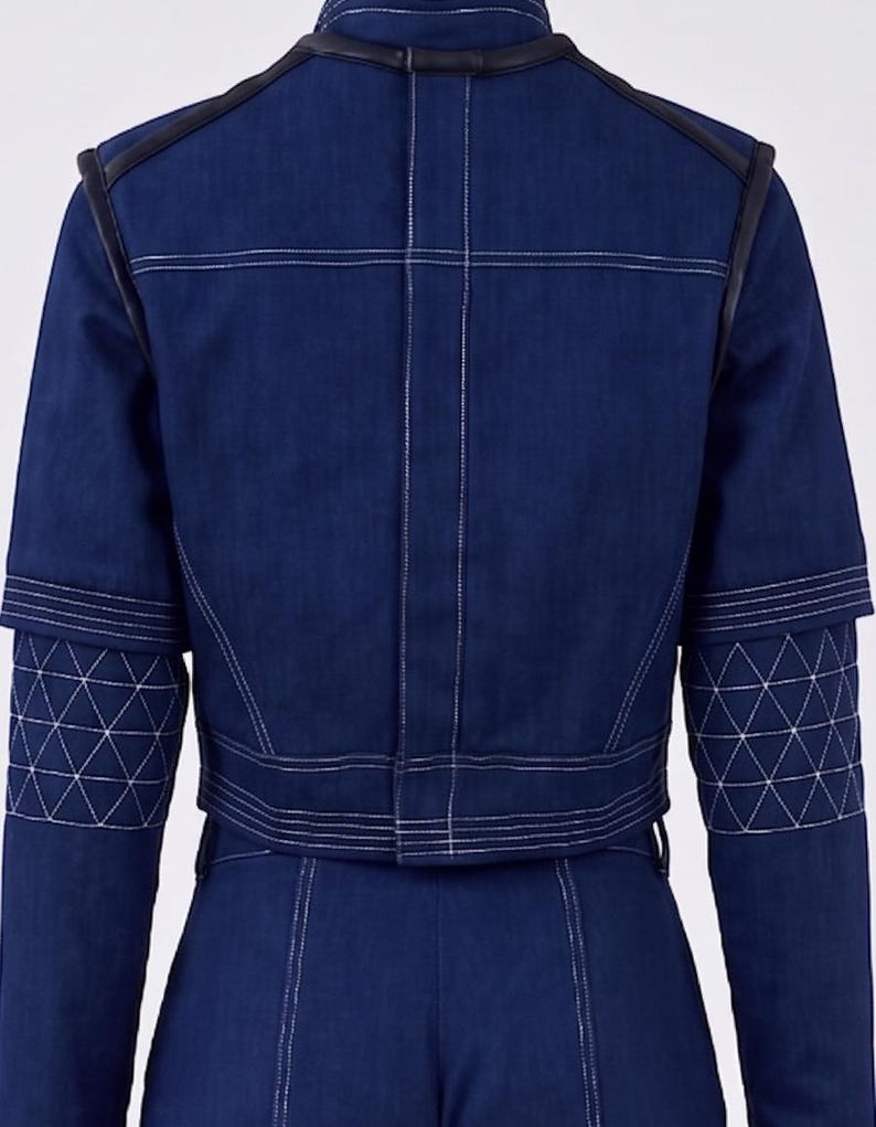 Op de afbeelding: Een donkerblauw spijkerjack met een opstaande kraag, zwarte lederachtige biezen op de schouders en witte contraststiksels. De mouwen hebben een gewatteerd driehoekspatroon en het jack is gecombineerd met een bijpassende donkerblauwe spijkerbroek.