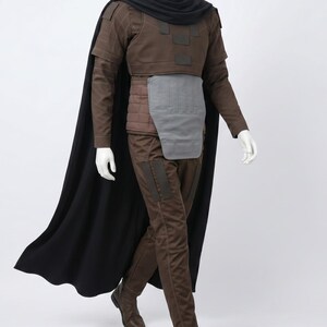 May include: A brown long-sleeved tunic with a high collar, a black cape, and matching brown trousers. The outfit includes a grey rectangular abdominal plate, a quilted brown waist section, and dark brown boots, all displayed on a white mannequin.