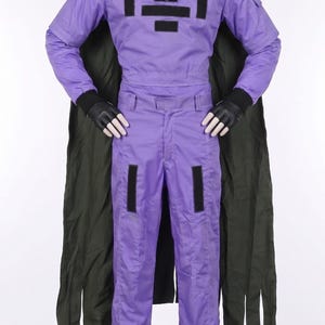 May include: A purple jumpsuit with long sleeves and a high collar, featuring black rectangular patches on the chest and thighs. The outfit includes a dark olive green cape draped over the shoulders and black gloves, worn on a white mannequin figure.