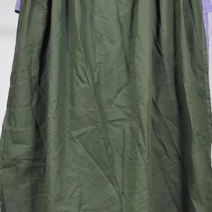 May include: A long, olive green cape featuring vertical pleats along the upper section and a series of rectangular cutouts along the bottom hem. The cape is worn over a light purple garment with a high collar and visible white stitching details.