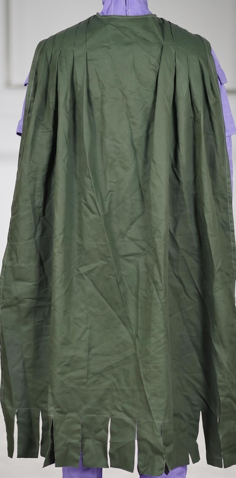 May include: A long, olive green cape featuring vertical pleats along the upper section and a series of rectangular cutouts along the bottom hem. The cape is worn over a light purple garment with a high collar and visible white stitching details.