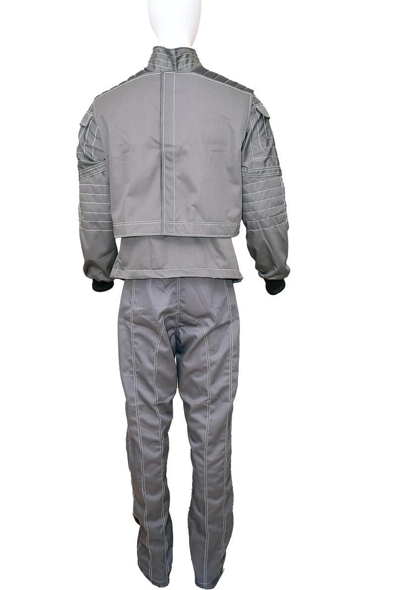 May include: A grey two-piece outfit consisting of a long-sleeved jacket and matching trousers. The jacket features a high collar, structured shoulder panels with horizontal stitching, and white contrast piping. The trousers have vertical seams.