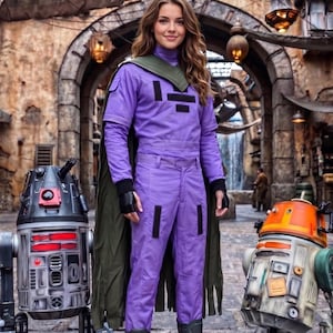 May include: A person wearing a purple long-sleeved jumpsuit with black rectangular accents, a dark green cape, and black boots, standing between a grey and red cylindrical droid and an orange and white cylindrical droid in a stone-walled area.