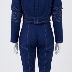 Op de afbeelding: Een donkerblauwe spijkerjumpsuit met een hoge kraag, lange mouwen met doorgestikte elleboogstukken en witte contraststiksels. Het kledingstuk heeft een aansluitend lijfje, een gedefinieerde taille en een broek met rechte pijpen.