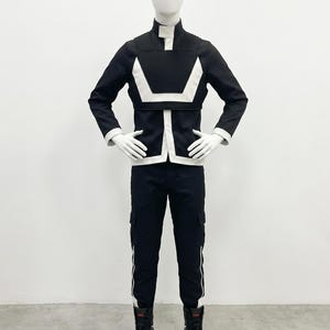 May include: A black and white long-sleeved jacket with a high collar and ribbed shoulder detailing, paired with matching black cargo-style trousers featuring white side stripes and black high-top sneakers worn on a white mannequin figure.
