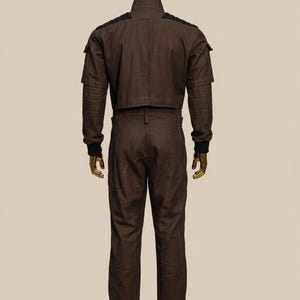 May include: A brown jumpsuit with a cropped jacket and matching pants. The jacket has a high collar and black accents on the shoulders. The pants have a straight leg design. The mannequin has a gold head.