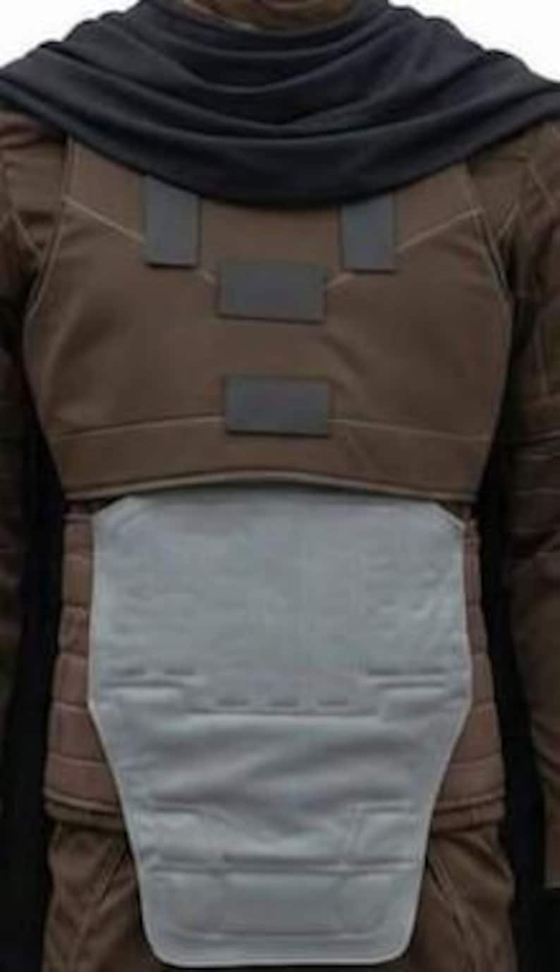 May include: A brown tactical vest featuring a dark grey cowl draped over the shoulders, a chest plate with three rectangular dark grey accents, and a large, light grey abdominal armour plate with moulded geometric detailing and horizontal stitching.