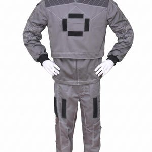 May include: A gray two-piece uniform set consisting of a long-sleeved jacket and matching trousers. The jacket features a high collar, dark gray shoulder panels, and a central square emblem made of four black rectangular patches with white stitching.