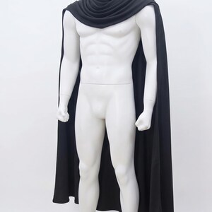 May include: A solid black cape featuring a draped, cowl-style neckline that rests around the shoulders. The fabric is smooth and opaque, extending down the back and sides in a long, flowing silhouette that reaches past the waistline.