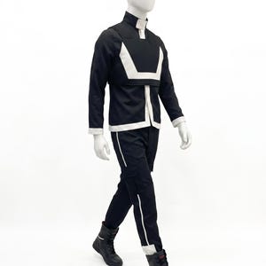 May include: A black long-sleeved jacket with a high collar and a white geometric chest panel, paired with matching black trousers featuring a white vertical stripe down the leg and black lace-up boots, displayed on a white mannequin.