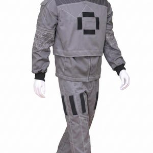 May include: A grey two-piece outfit consisting of a long-sleeved jacket and matching trousers. The jacket features a high collar, black rectangular accents on the chest, and quilted shoulder panels. The trousers have black rectangular patches.